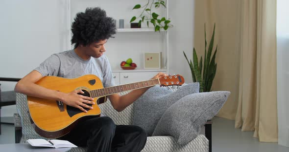 Afro American Boy Teenager Male Musician Black Man Artist Sitting on Sofa at Home Tuning Guitar