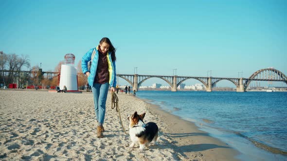 Young Woman Walking With Cute Dog Welsh Corgi Along Sandy Beach on Sea Shore