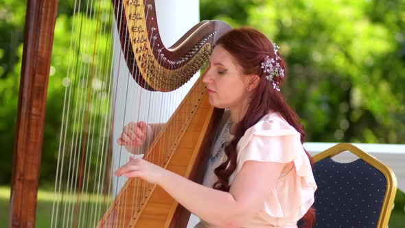 A woman with red hair plays a harp exquisitely in a flowery garden. Slow motion