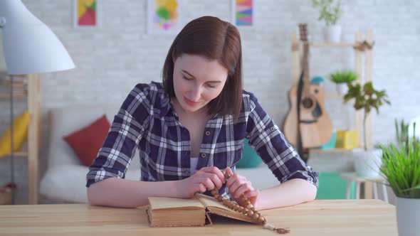 Portrait Young Woman Sitting at a Table with a Rosary in Her Hands and Reading a Book