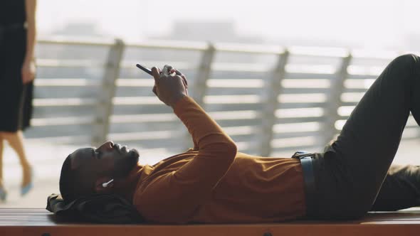 African American Man Lying on Bench and Using Smartphone