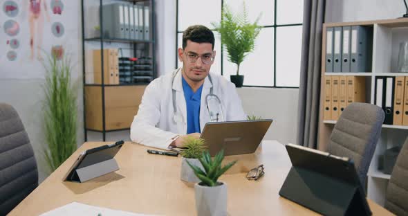 Male Doctor in Eyeglasses and white Coat Working on Laptop in Medical Office and Looking at camera