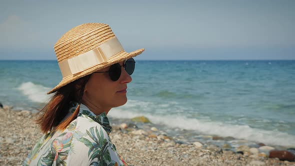 Portrait of cheerful carefree woman in straw hat sitting on beach and looking at horizon at hot summ