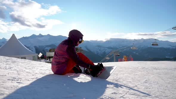 Girl with Snowboard Sitting on Top and Enjoying Mountain Landscape. Sport Woman in Snowy Mountains