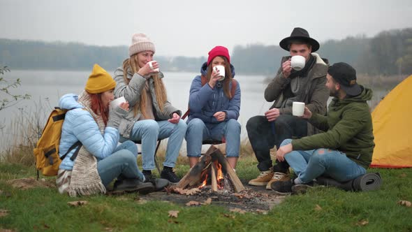 Joyful Young People Friends Are Clinking with Drinks Sitting Around Fire in Forest with Warm