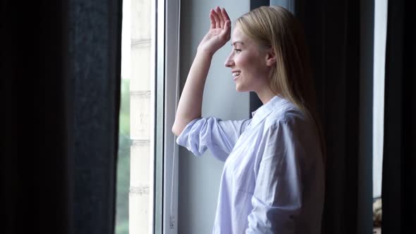 A pretty woman is standing by the window inside the apartment waving to someone and smiling