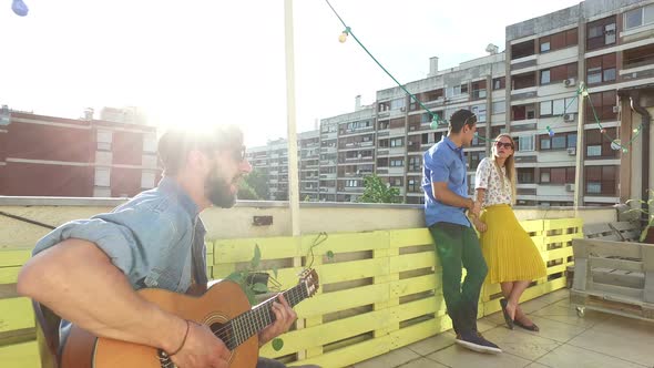 Musician playing guitar at rooftop party, people hanging out in background