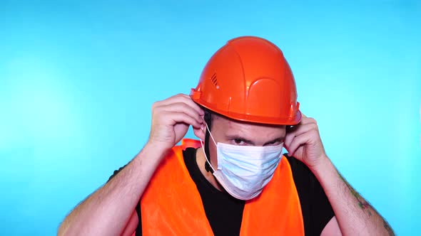 Male Construction Worker in Overalls Putting on Medical Mask on Face on Blue Background