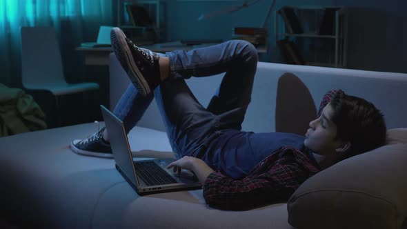 Male student relaxing on couch with laptop, watching images in social network