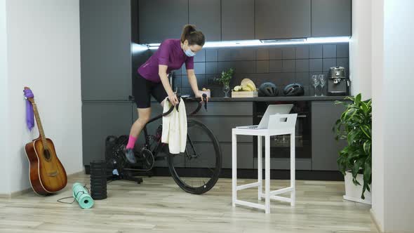 Fit caucasian female in protective medical mask training on smart bicycle trainer at home 