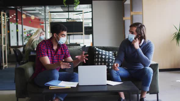 Diverse business people wearing masks using laptop and goign through paperwork in modern office