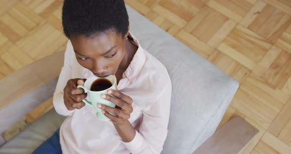 Happy african american woman sitting on armchair in living room, drinking coffee