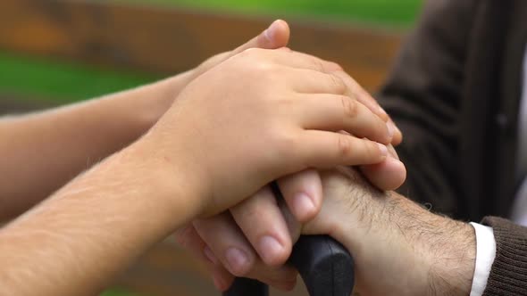 Hands of Boy Carefully Holding Hands of Old Man With Cane Sitting on Bench