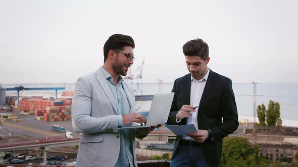 Two Business Partners Discussing the Meeting with Laptop and Contract Outdoors on Background of the