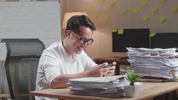 Asian Man Enjoys Playing Game On Smartphone After Working With Documents At The Office