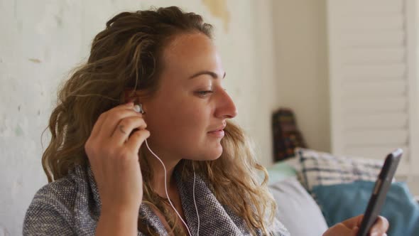 Happy caucasian woman wearing earphones sitting on sofa using smartphone in cottage living room