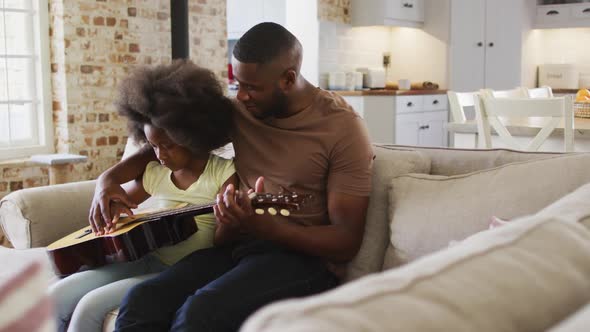 African american father and his daughter sitting on couch playing guitar together