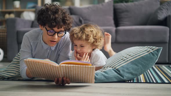 Young Woman Mommy Reading Book To Curious Boy Discussing Story on Floor at Home