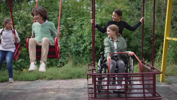 Front View Happy Disabled Girl in Wheelchair Swinging on Accessible Swings with Cheerful Friends