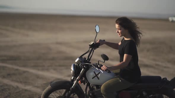 Beautiful Young Woman Riding an Old Cafe Racer Motorcycle on Desert at Sunset or Sunrise