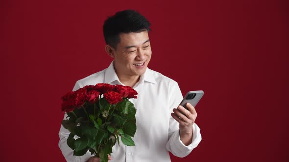 Positive Asian man holding a bouquet of red roses and typing by phone