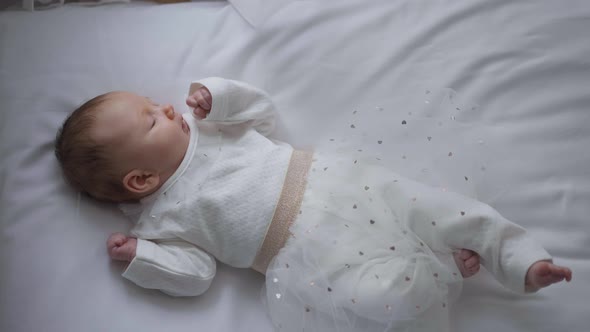 Top View Wide Shot Charming Newborn Baby Girl in White Dress Lying on Cozy Bed at Home Indoors