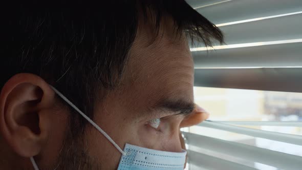 A man in a medical mask looks out the window through a slit in the blinds with sadness in his eyes