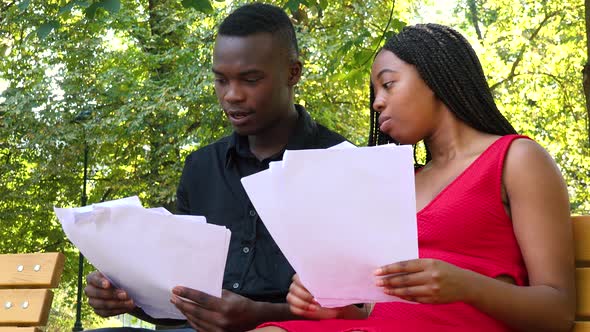 A Black Man and a Black Woman Sit on a Bench in a Park with Papers, the Man Explains Something