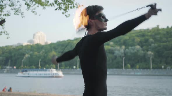 Young Slim Man in Black Clothes and Mask Performing a Show with Flame Standing on Riverbank