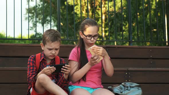 The Older Sister and Brother Are Sitting on the Street, Looking at Smartphones.