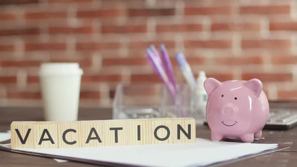 Hand Puts Money in Piggy Bank Next to Which There are Cubes with Word Vacation