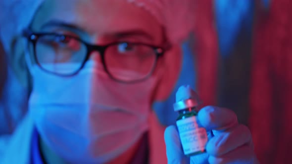 Male doctor in a protective mask shows a bottle of vaccine and recommends it in emergency light