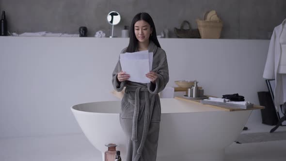 Front View of Smiling Asian Slim Young Woman in Bathrobe Standing in Bathroom Examining Paperwork