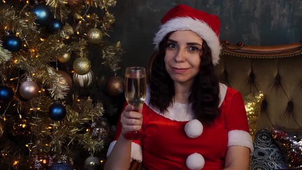 Young Woman in Santa Claus Costume with Glass of Champagne Sitting on Chair Near Christmas Tree