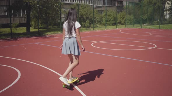 Young Girl in Sneakers, Skirt and T-shirt Riding Yellow Skateboard on an Outdoor Basketball Court