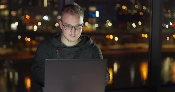 A young man works online on a laptop against the backdrop of a night city. 