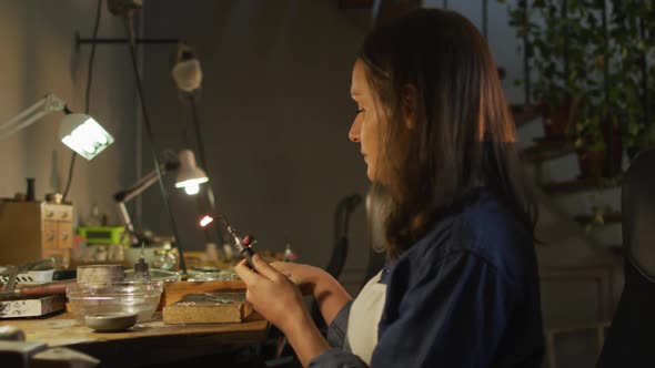 Focused caucasian female jeweller sitting at desk, making jewelry in workshop