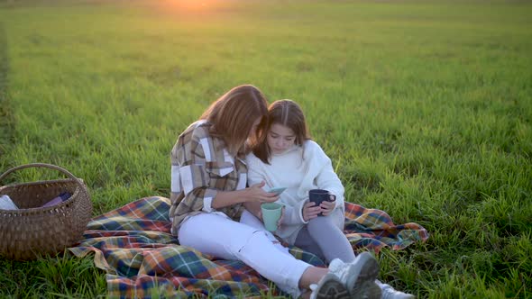 Young mum with a cute daughter use smartphone sit on plaid on a field at sunset