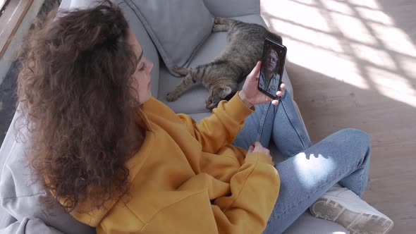 Young Hispanic Girl Sit on Sofa with Cat Hold Phone Video Call on Mobile Screen