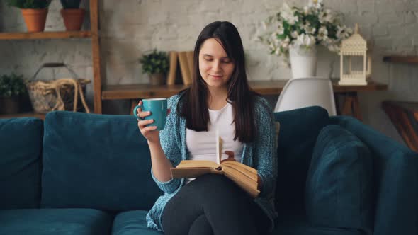 Girl Is Reading Book Sitting on Sofa at Home with Cup of Tea and Relaxing Enjoying