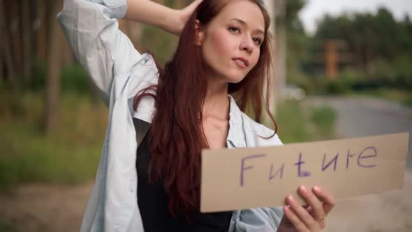 Gorgeous Redhead Young Woman with Future Banner Standing at Forest on Suburban Road Waiting for Car