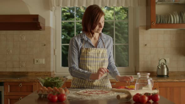 30s Woman in Apron Placing Ingredients for Pizza Standing in the Kitchen