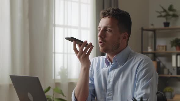 Millennial Attractive Man in Shirt Sit at Desk in Modern Home Office Holding Smartphone Communicates