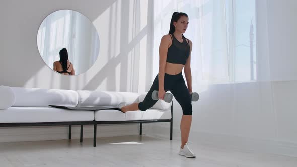 Brunette in Black Sportswear in a White Apartment Makes a Split Squat with Dumbbells in Her Hands