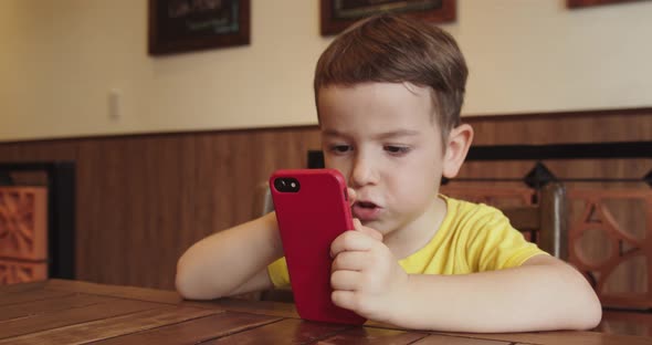 Portrait of a Little Boy Cute Child with a Looking at the Phone While Sitting at the Table a