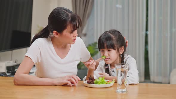 Asian Mother teach and motivate unhappy young girl child try to eat green vegetable on dinner plate.