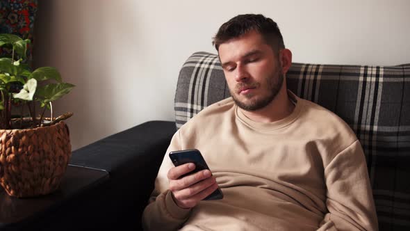 Bored Young Caucasian Man Scrolling Phone at Home, Reading Uninteresting Information