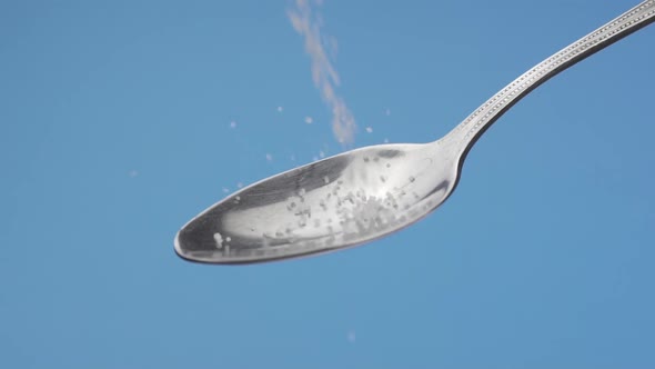White Sugar Slowly Fills a Teaspoon and Crumbles on a Blue Background