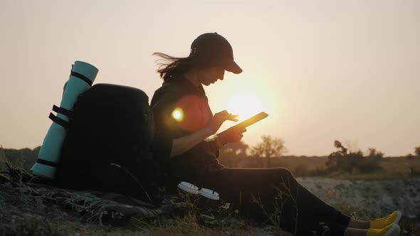 Hipster Young Girl with Backpack on the Top of a Rock Enjoys the View of Sunset and Using Digital