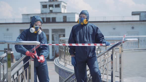 Two Caucasian People in Biohazard Suits Using Red and White Caution Tape on City Street. Young Man